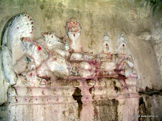 Lord Vishnu reclining on the coiled serpent called Sesha. The hood of the serpent makes a cover over Vishnu's head. You can also spot the symbols of Vishnu, the conch and the Chakta (the discus of Vishnu) near his hands. His first left arm supports his head and the second left arm holds the Chakra. The first right arm holds the Counch, called Panchajanya the other right hand rests on his body.

Seen in the middle above Vishnu is the creator god Brahma. The four headed Brahma sits on a lotus flower sprouted out of Vishnu's naval.

At the right extreme, sitting by the feet of Vishnu are his two consorts.

You can also spot the club of Vishnu near his feet.  

The whole image is carved on the vertical face of a large boulder. The sanctorum is built around this image, a technique used in many temples in Hampi.

This is in the temple called Rangatha Temple, located at the end of the Courtesan's Street by the riverside. Visit this as part of your riverside ruins itinerary.